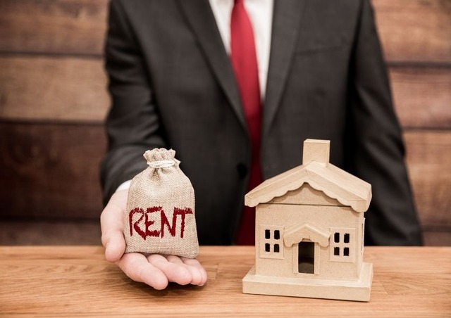 business-person-holding-a-bag-with-the-word-rent-on-it-next-to-a-wooden-house